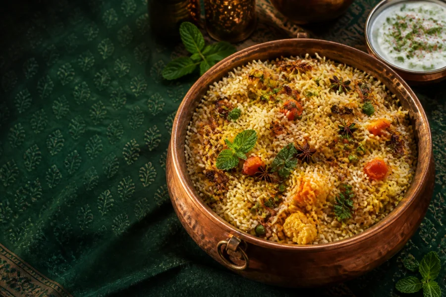 Vegetable biryani in a copper handi with mint and cucumber raita on green block-print cloth